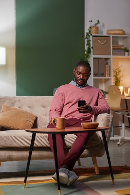 Afrikaanse jongeman zittend op de bank voor de tafel met een kopje koffie en zijn mobiele telefoon gebruiken tijdens de koffiepauze