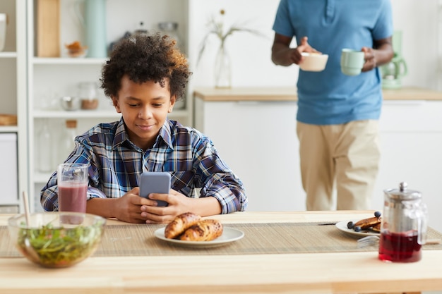 Afrikaanse jongen die aan de tafel zit en zijn mobiele telefoon gebruikt terwijl zijn vader het ontbijt in de keuken voorbereidt