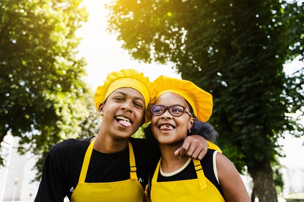 Afrikaanse kinderen koken in koksmuts en gele uniformen grimassen en tonen elkaars tong Afrikaanse tiener en zwart meisje hebben plezier en koken eten