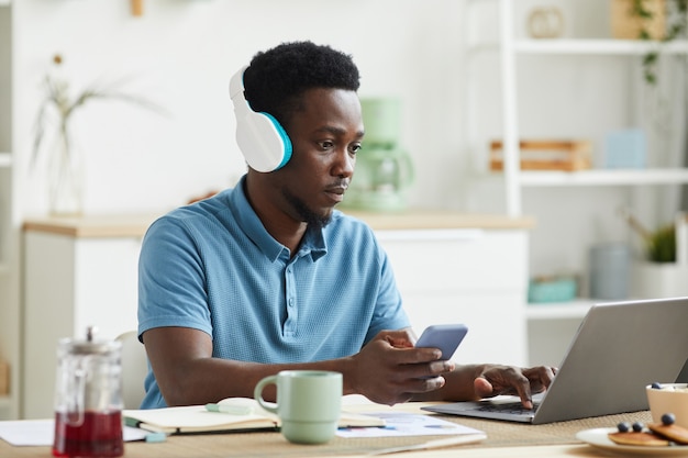 Afrikaanse man in koptelefoon concentreren op zijn online werk hij met behulp van laptop en mobiele telefoon terwijl hij thuis werkt