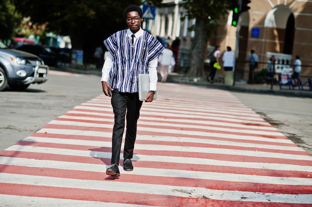 Afrikaanse man in traditionele kleding en glazen lopen op zebrapad met mobiele telefoon en laptop Afrika business.