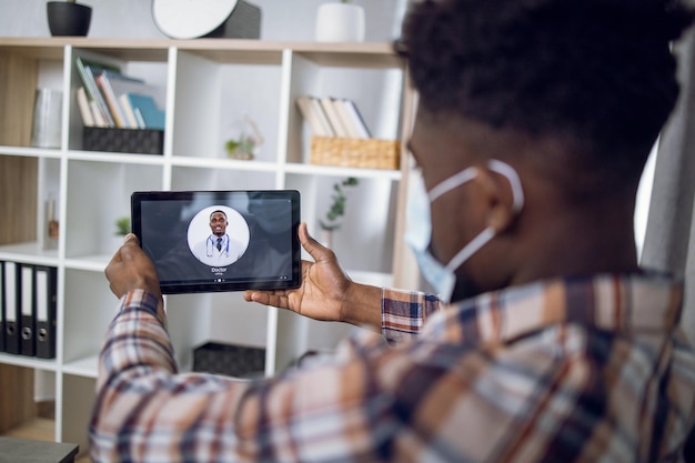 Afrikaanse man met gezichtsmasker die dokter belt op digitale tablet