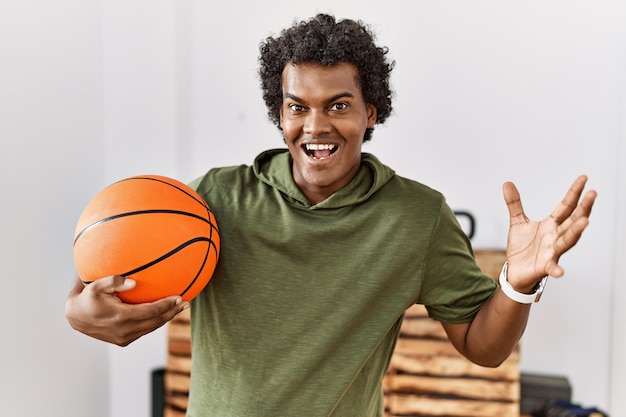 Afrikaanse man met krullend haar met basketbalbal in de sportschool die de overwinning viert met een gelukkige glimlach en winnaarsuitdrukking met opgeheven handen
