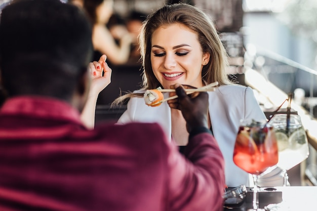 Afrikaanse man voedt zijn blonde vriendin sushi-broodjes op het zomerterras.