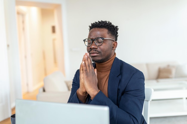 Afrikaanse man zit aan bureau met geklemde handpalmen gesloten ogen gebedshanden teken van hulp vragen