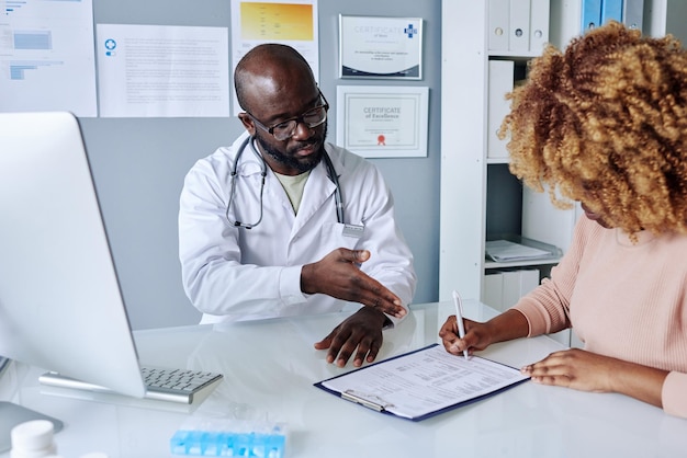Afrikaanse mannelijke arts in witte jas die zijn patiënt voorstelt om vóór de operatie een medische overeenkomst te ondertekenen