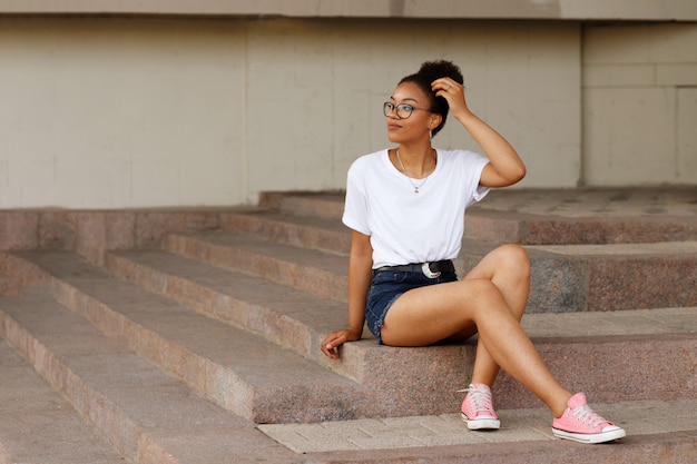 Afrikaanse meisje in een wit T-shirt en een bril zit op de trappen. Zomer