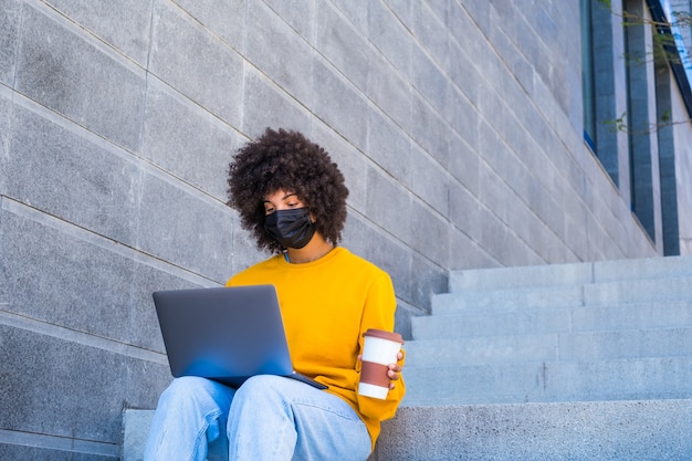 Afrikaanse of Amerikaanse zakenvrouw die alleen op straat werkt, zit op de trappen van de stad met haar laptop of computer met een masker op