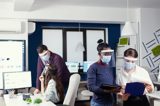 Afrikaanse ondernemer deelt creatieve ideeën en meningen met collega's die een gezichtsmasker dragen als veiligheidsmaatregel. Multi-etnisch zakelijk team dat werkt met respect voor sociale afstand tijdens wereldwijde pandemie met co