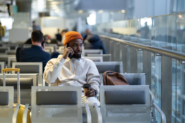 Afrikaanse reiziger die koffie drinkt en wacht op vliegen op de luchthaventerminal terwijl hij op mobiel praat