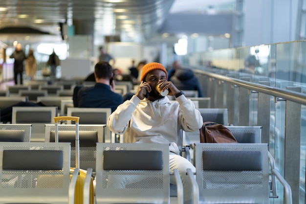 Afrikaanse reiziger die koffie drinkt en wacht op vliegen op de luchthaventerminal terwijl hij op mobiel praat