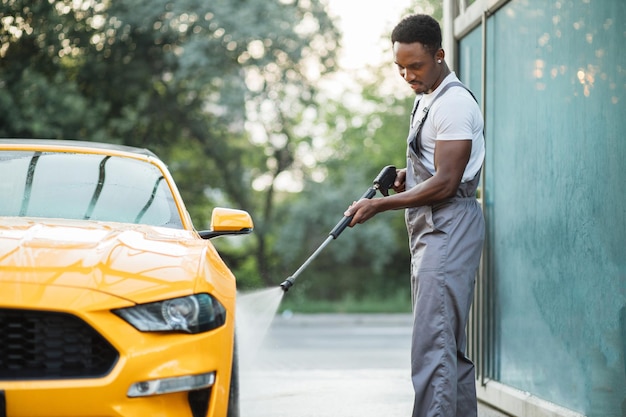 Afrikaanse stijlvolle jonge gefocuste man in werkkleding die de gele auto wast met een waterpistool