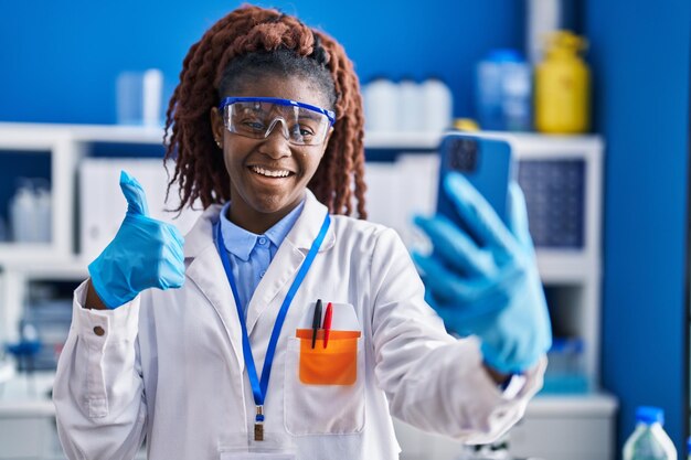 Afrikaanse vrouw die bij een wetenschapperlaboratorium werkt en een videogesprek doet, glimlachend, blij en positief, doet het uitstekend en goedkeuringsteken