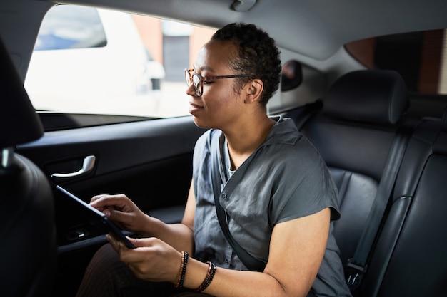 Afrikaanse vrouw die op de achterbank van de auto zit en online werkt met digitale tablet