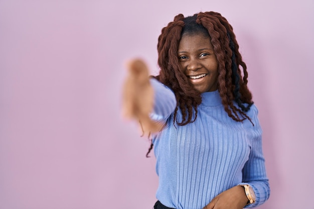 Afrikaanse vrouw die over een roze achtergrond staat en vriendelijk glimlacht en handdruk aanbiedt als begroeting en verwelkoming. succesvol bedrijf.