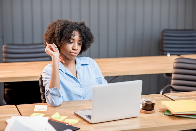 Afrikaanse vrouw die werkt met een laptop die aan het moderne kantoorinterieur zit