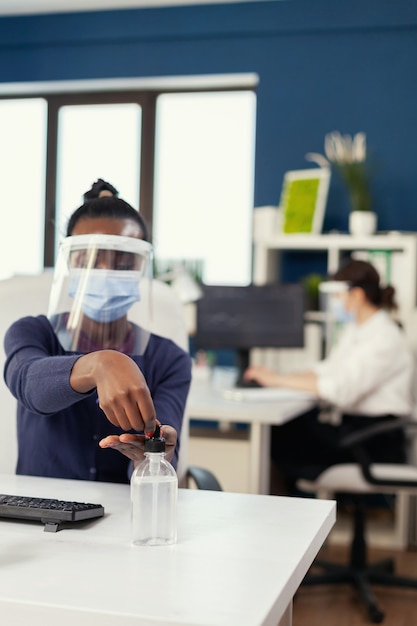 Afrikaanse vrouw in bedrijfskantoor die handdesinfecterend middel toepast met gezichtsmasker tijdens coronavirus. zakenvrouw in een nieuwe normale werkplek die desinfecteert terwijl collega's op de achtergrond werken.