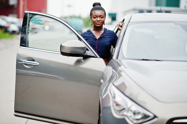 Afrikaanse vrouw in oranje broek en blauw shirt gesteld tegen zilveren suv auto met geopende deur