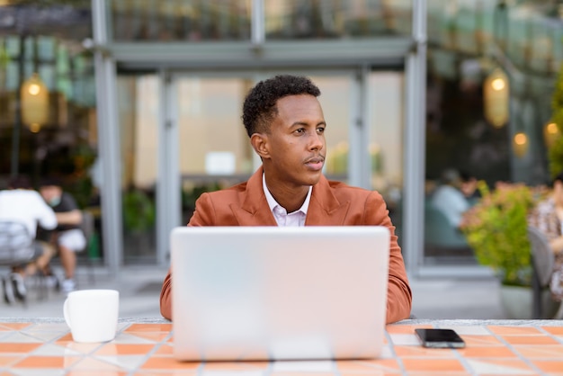 Afrikaanse zakenman buiten in coffeeshop met laptopcomputer terwijl hij denkt