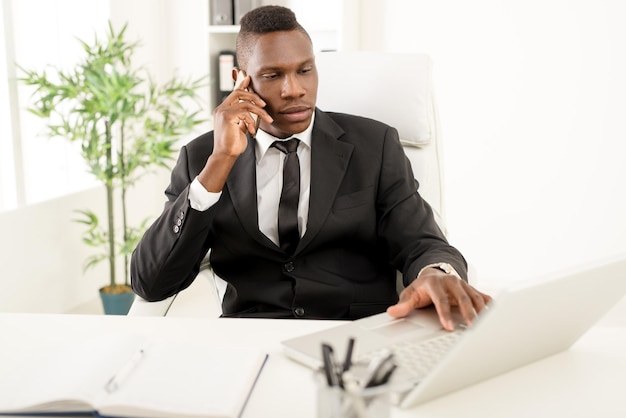 Afrikaanse zakenman die slimme telefoon gebruikt en aan laptop in het bureau werkt.