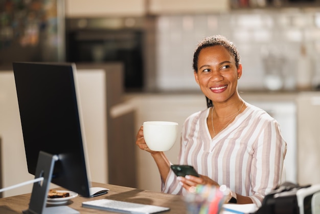 Afrikaanse zakenvrouw die een koffiepauze neemt en een mobiele telefoon gebruikt terwijl ze thuis werkt.