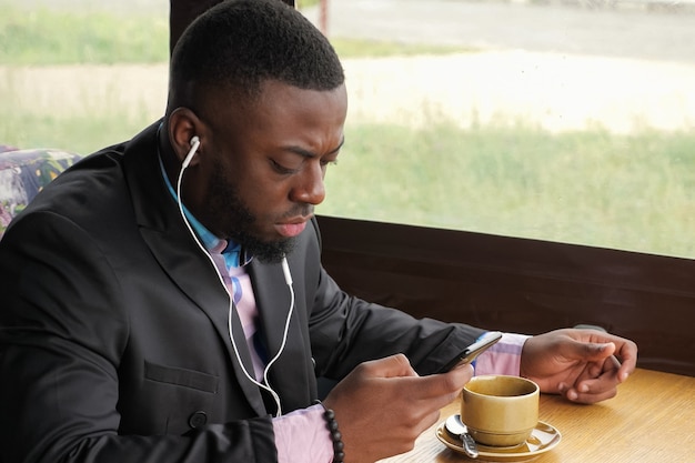 Afro american businessman in cafe. Black man is listening music in earphones browsing mobile phone and drinking coffee sitting in cafe. Guy is wearing in shirt and suit jacket. Enjoying morning time.