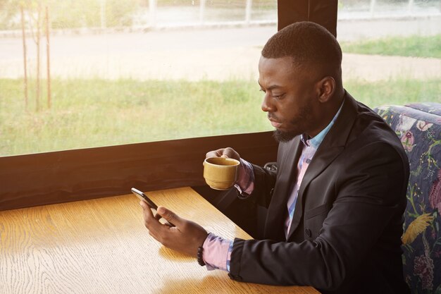 アフリカ系アメリカ人のビジネスマンは、カフェに座ってスマートフォンを閲覧しています。彼はメッセンジャーでチャットしているメッセージを入力しています。黒人の男は携帯電話、日光のソーシャルメディアでインターネットページをオンラインでめくっています