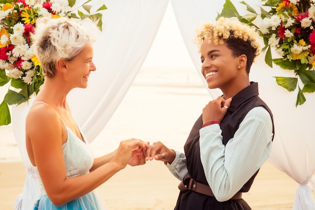 Afro american female in the role of the groom and short blonde haired bride in blue dress in ceremony under wedding arch in tropical beach