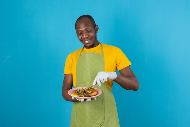青い壁に食べ物のプレートを保持している緑のエプロンのアフロアメリカ人