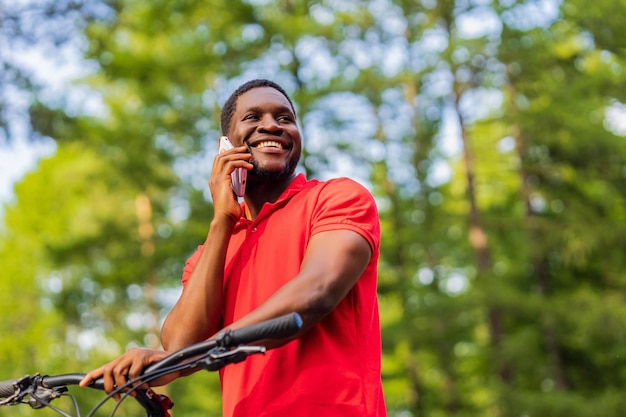 Uomo afroamericano in maglietta rossa che cammina nel parco con una bicicletta