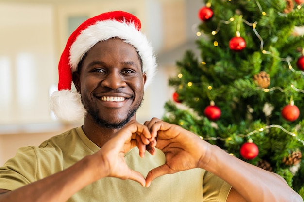 Афроамериканец показывает сердечный жест руками в шляпе Санты возле рождественской елки дома