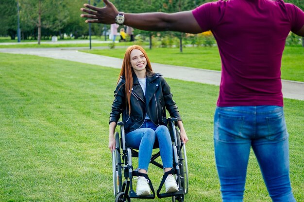 Foto uomo afroamericano seduto su sedia a rotelle, la sua ragazza rossa dai capelli rossi che rotola il passeggino nel parco autunnale. avere un appuntamento romantico.