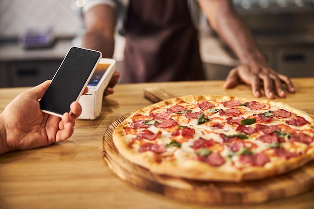 Photo afro american man standing at the counter with freshly baked pizza and using terminal for contactless payment while customer holding mobile phone