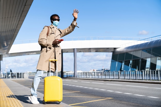 黄色いスーツケースを持ったアフリカ系アメリカ人の千年紀の男性が空港ターミナルに立ち、電話を使い、タクシーを呼び、手を上げ、インフルエンザウイルスとの接触から身を守るために顔の医療用マスクを着用します。covid-19