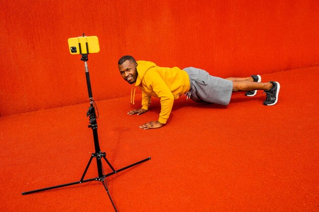 Afro american sporty man look at phone camera smiling while doing plank exercise practicing work out using webcam on smartphone on tripod positive active afro american do training online isolated