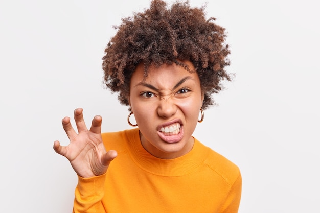 Afro American woman makes grimace scary face holds hand as if claw clenches teeth wants to scare child tells horror story wears casual clothes isolated over white  wall growls like animal