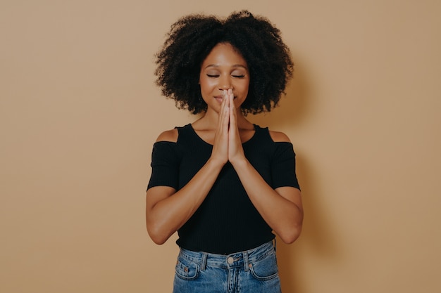 Afro American young woman praying and holding hands in prayer gesture standing isolated on dark beige studio background with copy space, smiling with eyes closed, Praying and hope concept