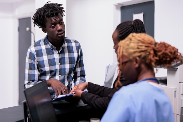 Afro-amerikaans medisch team in gesprek met volwassene bij de receptie van het ziekenhuis, rapportpapieren invullen voor overlegafspraak. patiënt die verzekeringssteun ontvangt van verpleegkundige en werknemer.