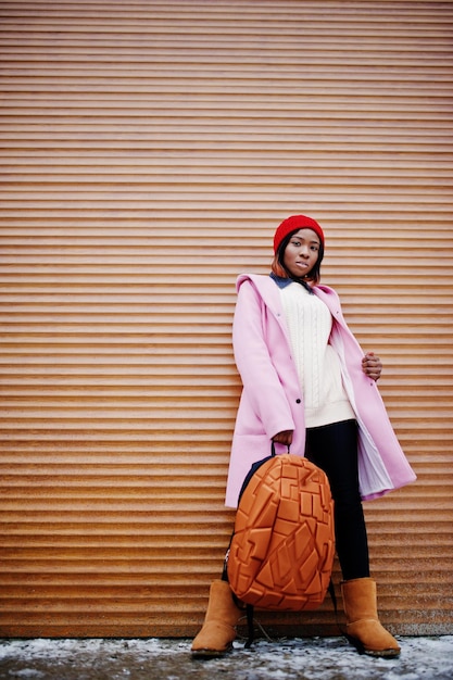 Afro-Amerikaans meisje in rode hoed en roze jas met rugzak tegen oranje luiken
