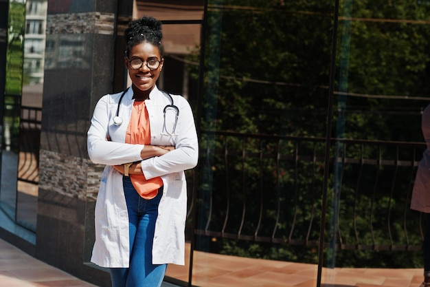 Afro-Amerikaanse arts vrouw bij laboratoriumjas bril met stethoscoop poseerde buiten tegen kliniek