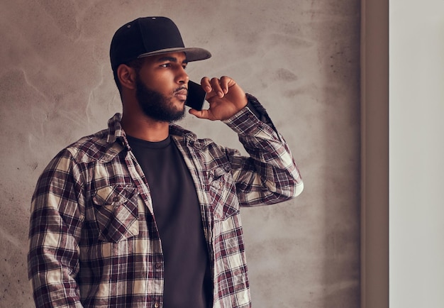 Afro-Amerikaanse bebaarde man met een geruit hemd en pet die telefonisch praat en naast een grijze getextureerde muur staat.