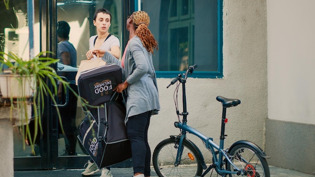 Afro-Amerikaanse bezorger die een papieren zakbestelling geeft aan de klant en een maaltijdpakket voor een restaurant op de fiets aflevert. Vrouw ontvangt afhaalmaaltijden fastfood van koerier, zittend bij de ingang van het gebouw.