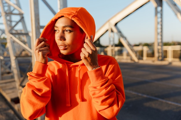 Afro-Amerikaanse blije vrouw in hoodie die oordoppen gebruikt tijdens het trainen op de oude brug