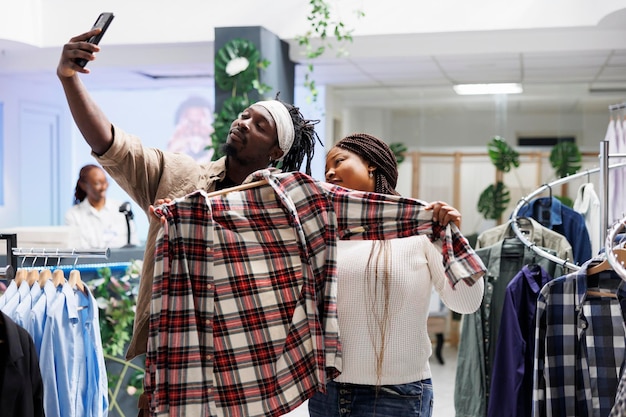 Afro-amerikaanse bloggers houden shirt vast en nemen smartphone-selfie om reclame te maken voor het modemerk in de winkel. Marketingspecialist die foto's van mobiele telefoons maakt voor een boetiek-account voor sociale media
