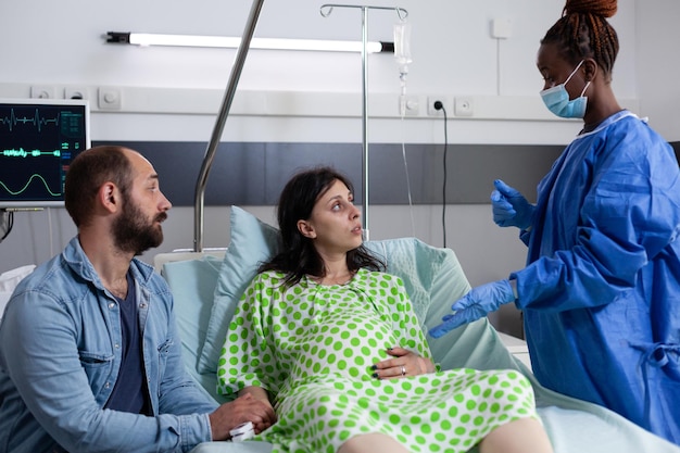 Afro-Amerikaanse chirurg die zwangere vrouw medische ondersteuning geeft en de bevallingsoperatie in de ziekenhuisafdeling uitlegt. Patiënt met zwangerschap die in bed ligt en contracties heeft voordat hij in de bevalling komt