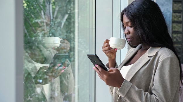 Afro-Amerikaanse dame drinkt koffie en sms't aan de telefoon