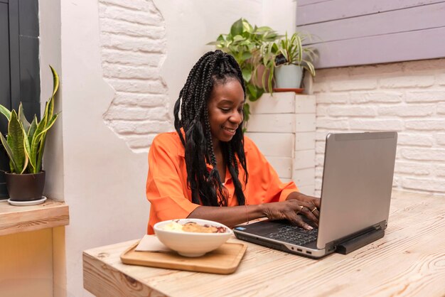 Afro-amerikaanse digitale nomade vrouwelijke ondernemer die met laptop werkt en op mobiele telefoon praat terwijl ze een gezond acai bowl-ontbijt heeft