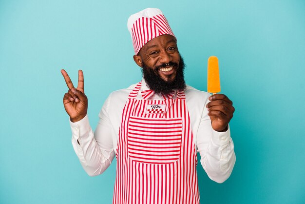 Afro-Amerikaanse ijsmaker man met een ijsje geïsoleerd op blauwe muur vrolijk en zorgeloos met een vredessymbool met vingers.