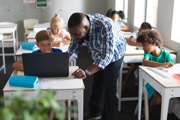 Afro-Amerikaanse jonge mannelijke leraar die laptop laat zien aan een blanke basisschooljongen aan een bureau