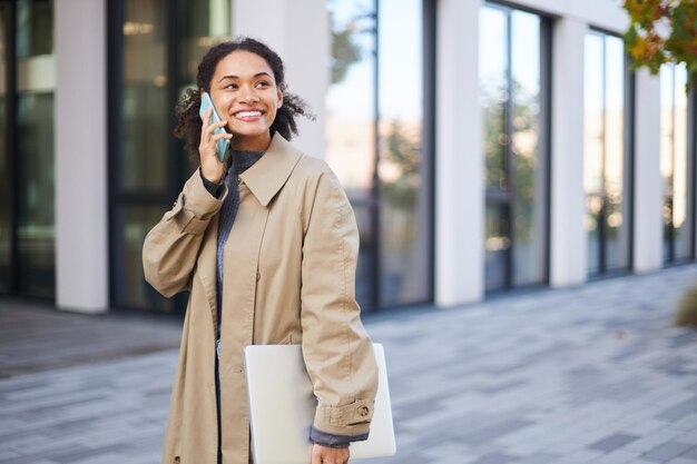 Afro-Amerikaanse jonge vrouw in beige casual jas die aan de telefoon praat terwijl ze door de straat slentert